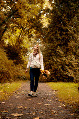 Obraz premium beautiful smiling girl in pink sport jacket walking in autumn park with a bouquet of autumn leaves holding in her hands