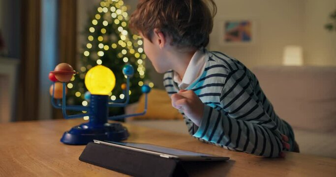 A 7-year-old boy with brown hair looks at pictures of planets on tablet and shows them on children's model of the solar system. He is wearing striped sweater. A Christmas tree flickers behind.