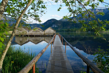 a pier leading to the wooden boat houses on tranquil lake Kochel (Kochelsee) with the scenic Bavarian Alps in the background reflected in the water (Bavaria, Germany)	