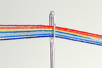 multi-colored threads for sewing in the form of a rainbow pass through an antique needle on a white background