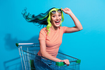 Photo of lovely young woman with vivid turquoise hair have fun riding in shopping cart isolated on...