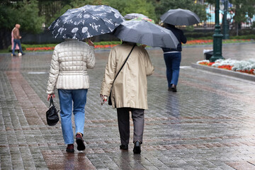 Rain in city, people with umbrellas walking on a street, two women in foreground. Rainy weather in autumn park