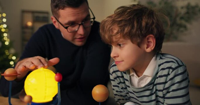 Close-up of father and son watching the rotation of the planets in model of the solar system. A young father with glasses explains son and shows the planets. The boy listens intently and smiles.