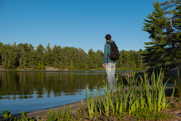 Young fit man in a black cap and hoodie with a backpack standing alone on the rocky shore of a lake...