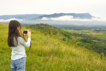 Back of an Asian girl who is standing and taking a view on her digital camera with the background of mountains and clouds.
