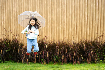 An Asian kid holding an umbrella is jumping over the grasses with brown grasses and a wooden wall in background.  A happy Asian kid with copy space from Thailand.