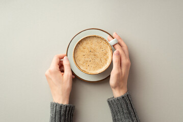 Autumn concept. First person top view photo of girl's hands in sweater holding cup of fresh frothy coffee on saucer on isolated grey background