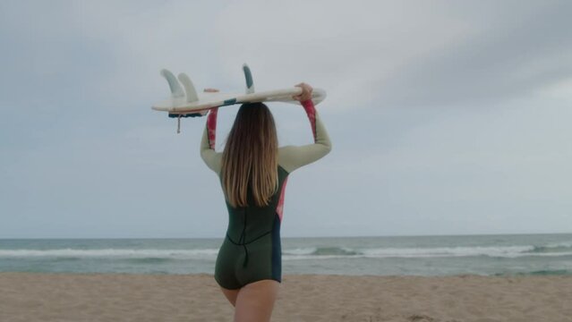 Attractive young woman in short wet suit carry surfboard on top of her head towards water. Surfing in the ocean on cloudy day. Cinematic female surfing