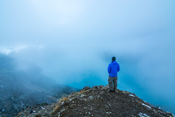 Fototapeta premium Adventurous athletic male hiker standing at the summit of Panorama Ridge looking down at Garibaldi Lake on a cloudy day. 