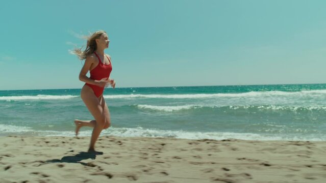 Side tracking shot of fit, athletic and beautiful young woman in red swimsuit run on empty beach in sunshine. Summertime bay coastguard watch concept. Healthy lifestyle of fitness lovers