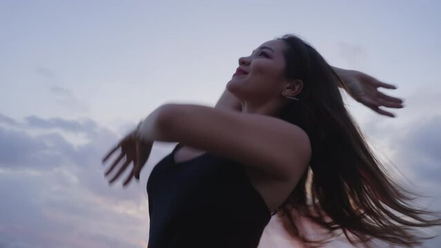 Slow motion footage of beautiful Korean girl wearing black silk evening dress dancing on a beach at sunset. Dynamic dance of woman with white skin at golden hour at seaside. Female moving with music.