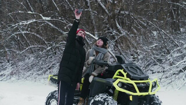 Young couple posing with ATV Quad bike in winter forest. Young man with a red hat takes selfie with his girlfriend on atv