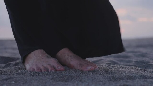Slow motion close-up view of female feet covered by sand under long maxi skirt on a beach at twilight. Close up footage of anonymous girl wearing winding silk evening dress on seaside after sunset.