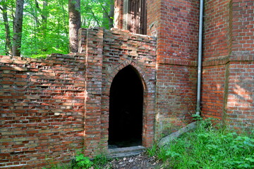 A close up on an  entrance to an old abandoned castle or church made out of red brick with the wooden door missing and the entire object being overgrown with vines, herbs, shrubs, and other flora