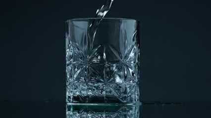 Pouring water crystal glass at dark background closeup. Liquid stream falling