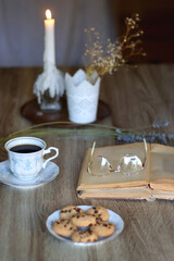 Cup of tea, plate with chocolate chip cookies, open book, reading glasses, lit candles and dry lavender flowers. Hygge at home, selective focus.