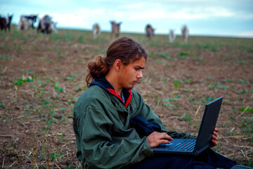 A teenage boy with a laptop grazes goats in a field. A goat herder in a field with a laptop communicates over the Internet. The concept of education, technology, survival.