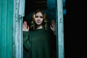 A beautiful mysterious blonde woman in a vintage green sweater stands outside the window in the darkness of an abandoned old creepy village house . Horror and mysticism.
