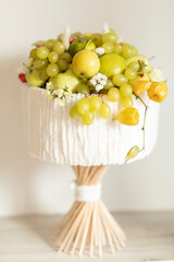 Ready isolated creative delicious edible handmade fruits bouquet, made of fresh food: grapes, apples, lemons, lime, physalis, flower roses, placed on bamboo skewers, stands on wooden table. Vertical