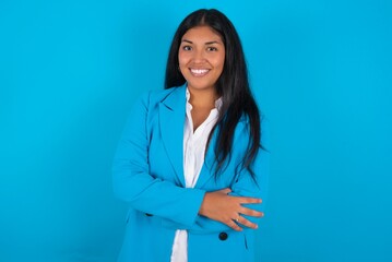 Young latin woman wearing  blue blazer blue background happy face smiling with crossed arms looking at the camera. Positive person.