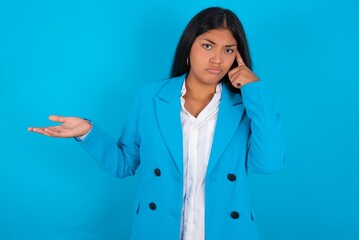 Young latin woman wearing  blue blazer blue background confused and annoyed with open palm showing...