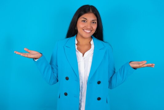 Cheerful cheery optimistic Young latin woman wearing  blue blazer blue background holding two palms copy space