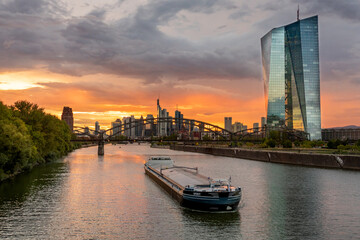Europäische Zentralbank (EZB) beim Sonnenuntergang mit dunklem Himmel mi Frankfurts Skyline im Hintergrund	