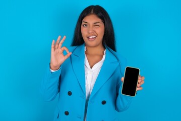 Young latin woman wearing  blue blazer blue background holding in hands cell showing ok-sign