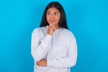 Thoughtful Young latin woman wearing gray sweater blue background holds chin and looks away pensively makes up great plan