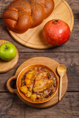 Chelnt with chicken in a wooden plate with a wooden spoon on the festive table for Rosh Hashanah next to apples, challah and pomegranate.