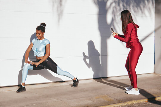 Young sportswoman photographing ethnic female friend exercising against white wall