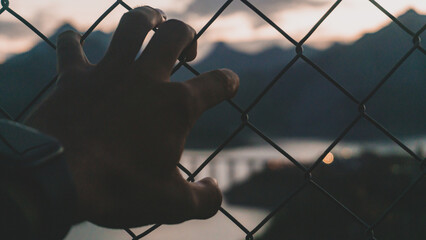 silhouette of a hand grabing a fence with the mountains at the bottom at sunset
