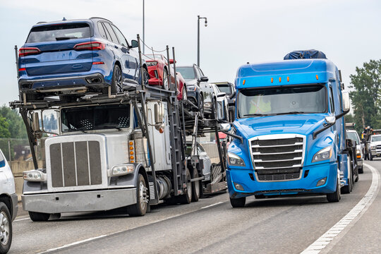 Heavy highway traffic with transporting cars white car hauler big rig semi truck driving side by side with blue semi truck transporting cargo on flat bed semi trailer