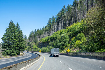 Industrial big rig semi truck transporting frozen cargo in reefer semi trailer running on the divided highway road with forest on the mountain in Columbia Gorge area