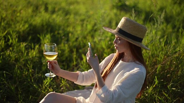 Pretty red-haired young woman blogger in straw hat taking photo of glass wine against beautiful landscape in sunny summer day, sitting on field among tall green grass. Shooting in slow motion.