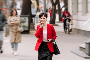 Young confident businesswoman in formalwear, red jacket is talking on smartphone while walking on city street. Telecommunication and mobile network for business concept. Female Business Style