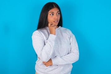 Fototapeta premium Astonished Young latin woman wearing gray sweater blue background looks aside surprisingly with opened mouth.