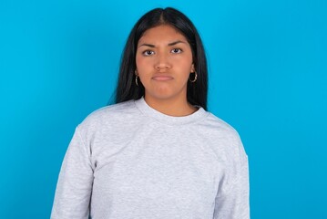 Displeased Young latin woman wearing gray sweater blue background frowns face feels unhappy has...