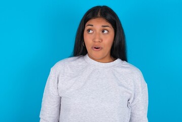 Shocked Young latin woman wearing gray sweater blue background look empty space with open mouth screaming: Oh My God! I can't believe this.