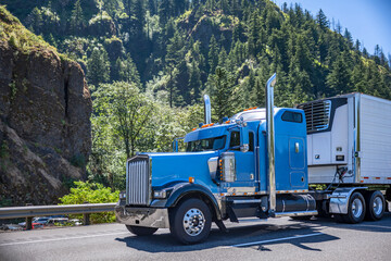 Shiny blue classic big rig semi truck tractor with reefer semi trailer transporting cargo running on scenic highway road with forest on the rocky mountain in Columbia Gorge National recreation area