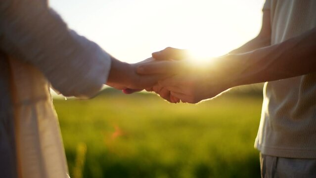 Close-up of unrecognizable young couple in love standing on field in front of each other, holding hands, stroking palms with fingers, on background of evening sunlight during beautiful summer sunset.