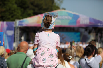 Obraz premium A happy child sitting on the shoulders of a parent standing in the crowd watching the performance of artists on the stage outdoors on a summer day