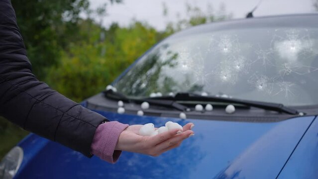 large hail in hand against the background of a car damaged in a storm