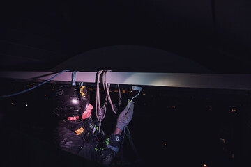 Industry night city work concept. Industrial mountaineering worker in uniform hangs over building...