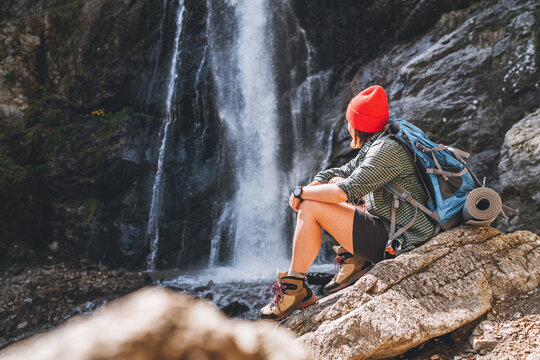 Woman with a backpack in red hat dressed in active trekking clothes sitting near the mountain river waterfall and enjoying the splashing Nature power. Traveling, trekking, and a nature concept image.