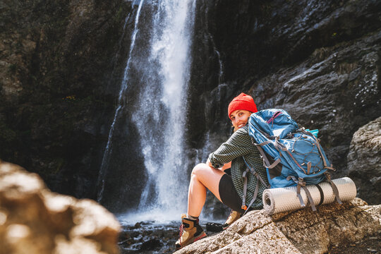 Smiling woman with backpack in red hat dressed in active trekking clothes and boots sitting near mountain river waterfall and enjoying splashing Nature power. Traveling, trekking, nature concept image