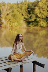 Fototapeta premium A young happy woman with a hippie smile sits on the lake shore on a bridge wearing eco clothing made of natural materials in harmony with nature in the fall. Travel lifestyle