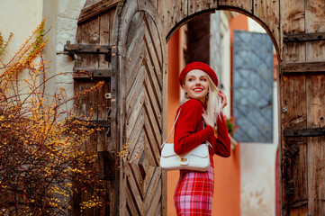 Fashionable happy smiling woman wearing stylish autumn outfit: red beret, cashmere sweater, checkered skirt, with trendy white bag, posing in street of European city. Copy, empty space for text 