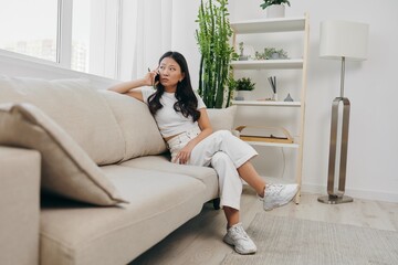 A young Asian woman sits at home on the couch relaxing and talking on the phone on her day off. Lifestyle without work in a comfortable home