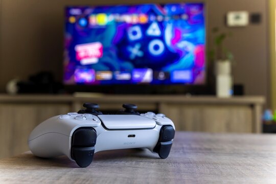 BRECHT, BELGIUM – AUGUST 14 2022: A portrait of a Sony Playstation 5 controller on a wooden table in front of a television showing the PS5 playstation plus screen in a living room.